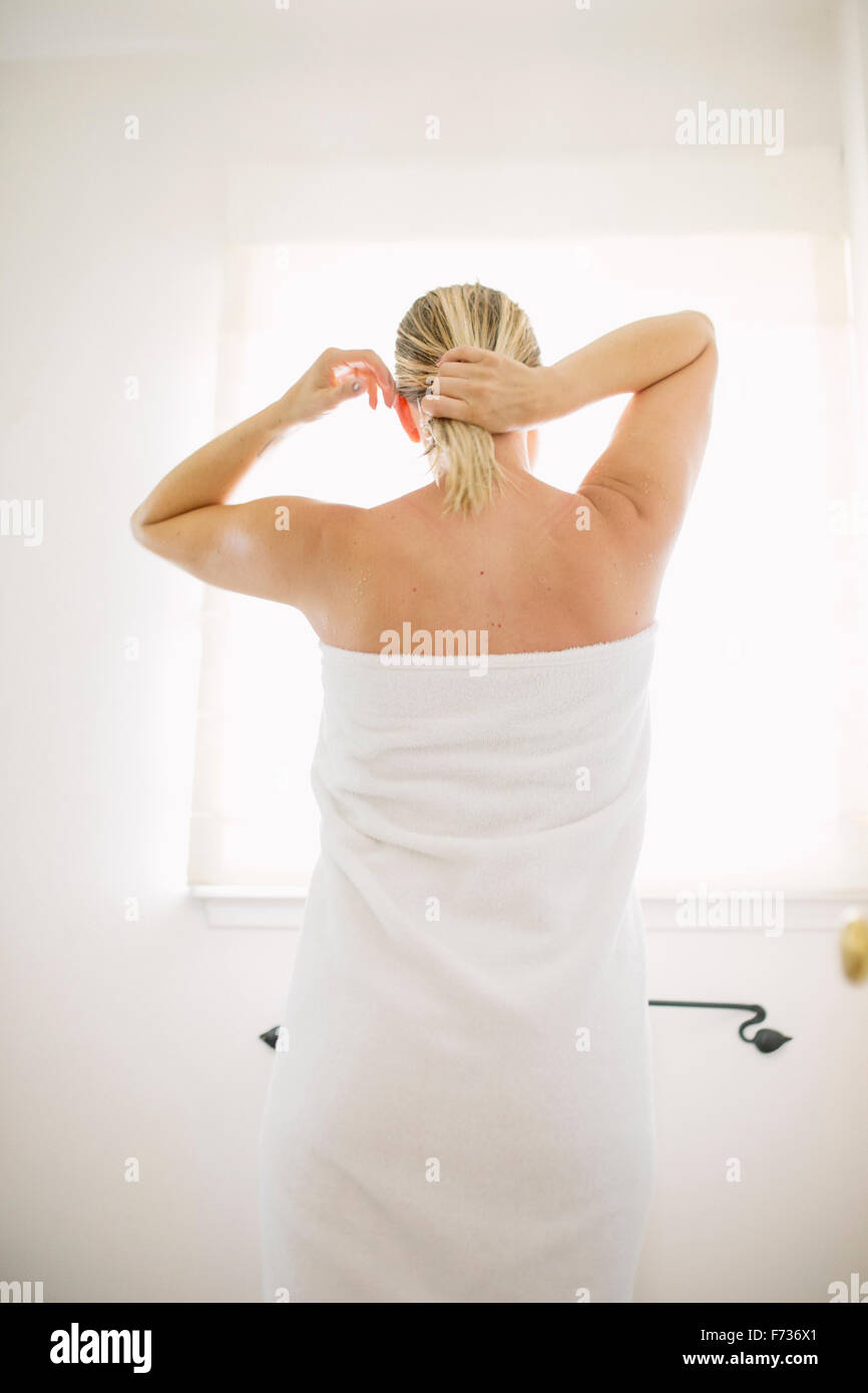 Mujer envuelta en una toalla blanca de pie en un baño, atando su cabello mojado. Foto de stock