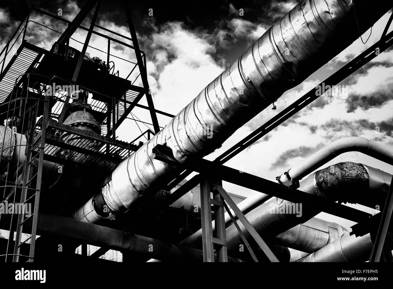 Tuberías de metal industrial de una fábrica que transporta agua/gas/materiales y pasarelas metálicas con un cielo nublado en blanco y negro mo Foto de stock