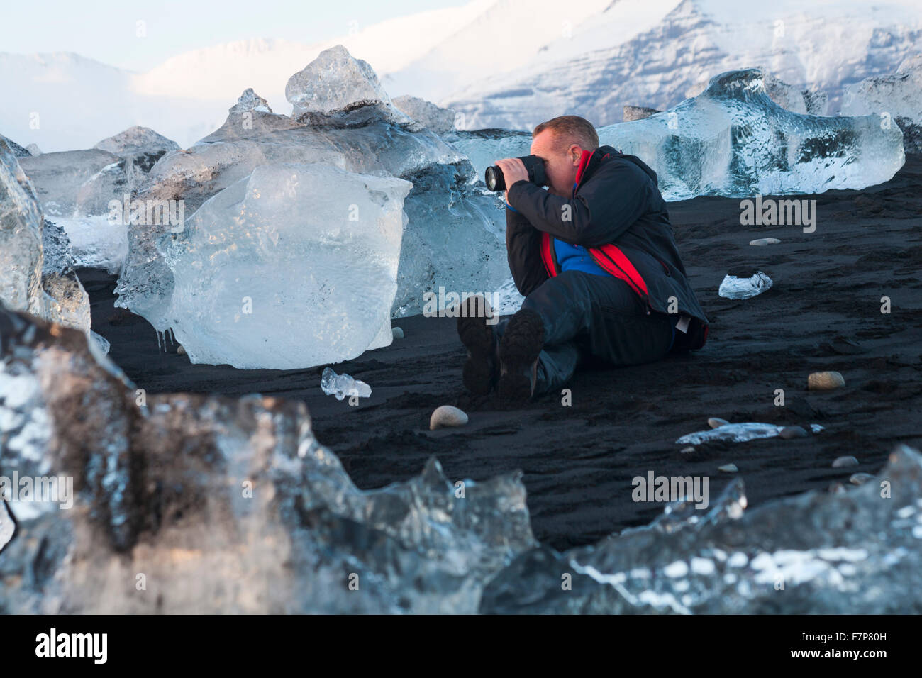 Fotógrafo con bloques de hielo en la playa en playa glacial Jokulsarlon, Diamond Beach, con montañas cubiertas de nieve en la distancia, Islandia en febrero Foto de stock