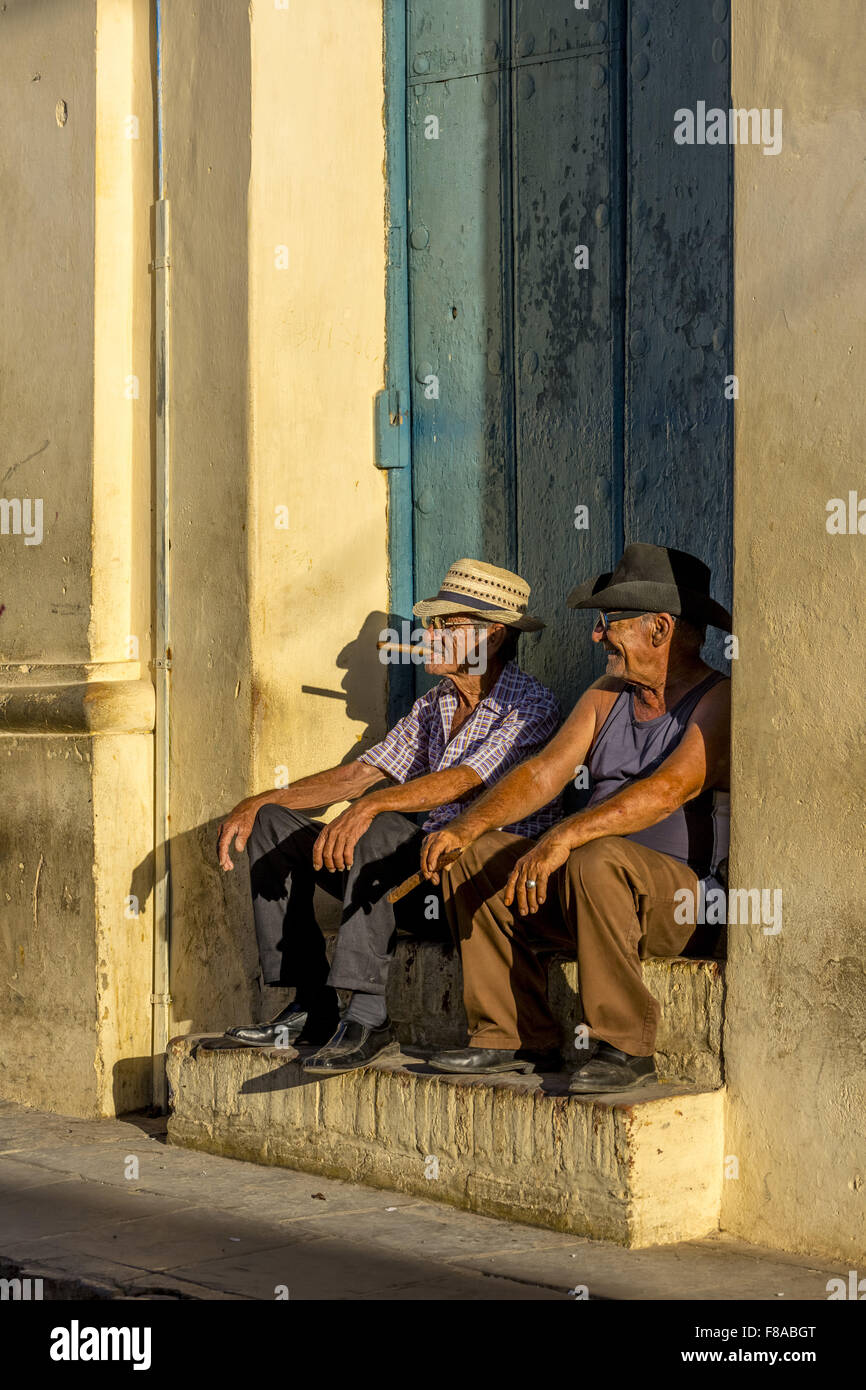 Dos viejos cubanos fuman cigarros y sentado en una escalera en la cálida luz del atardecer, Trinidad, Sancti Spíritus, Cuba, el Caribe, Foto de stock