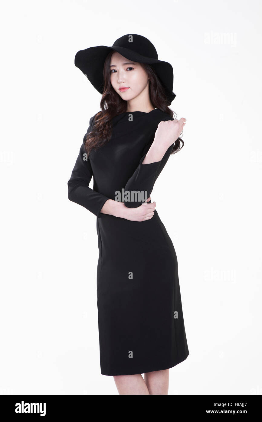 Mujer joven en un sombrero negro y vestido negro haciendo una pose con sus manos sobre su cintura y el otro brazo doblado hacia arriba Foto de stock