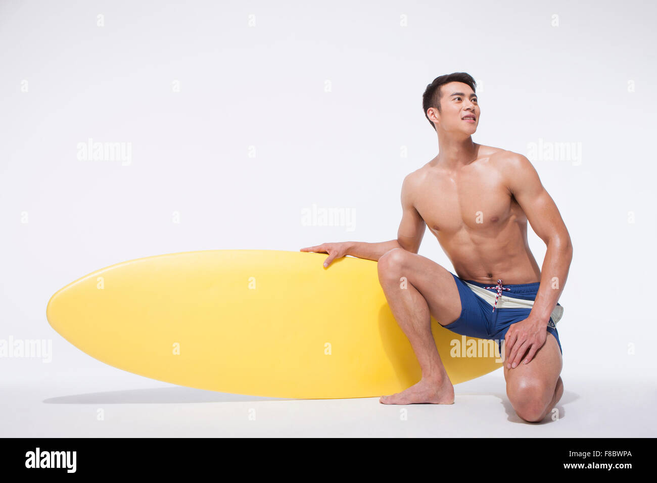El hombre muscular en natación pantalones de rodillas y la celebración de una junta de surf en el suelo y mirando hacia arriba Foto de stock