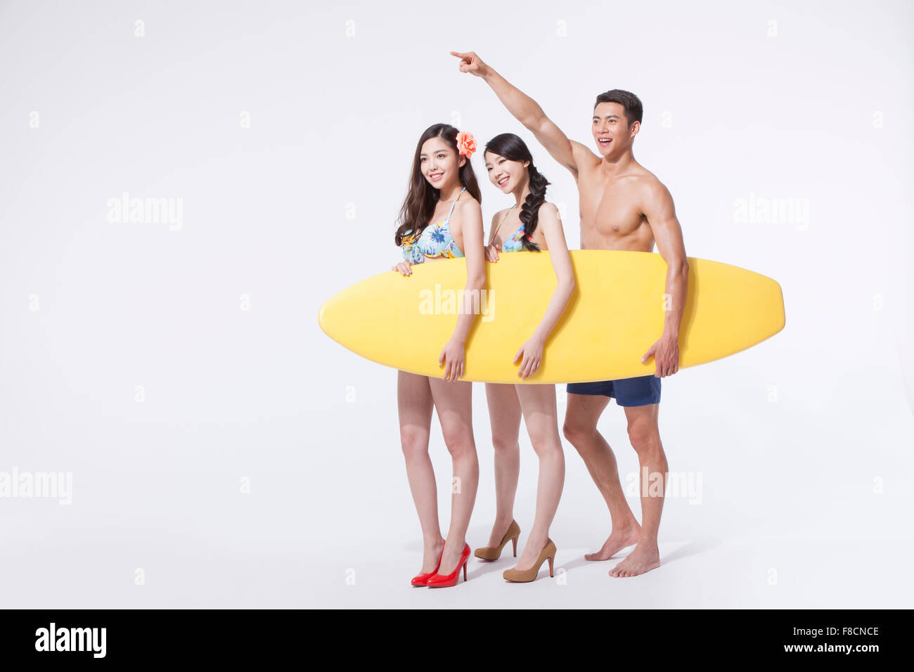 Dos mujeres en bikini y tacones y un hombre en natación pantalones la celebración de una junta de surf juntos y el hombre apuntando con el dedo. Foto de stock