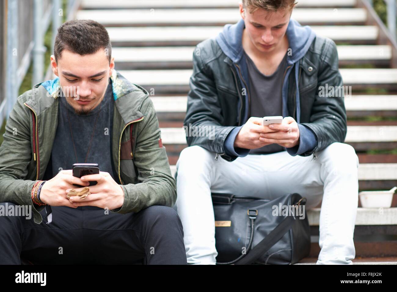 Dos hombres jóvenes, sentados en pasos, utilizando smartphones, al aire libre Foto de stock