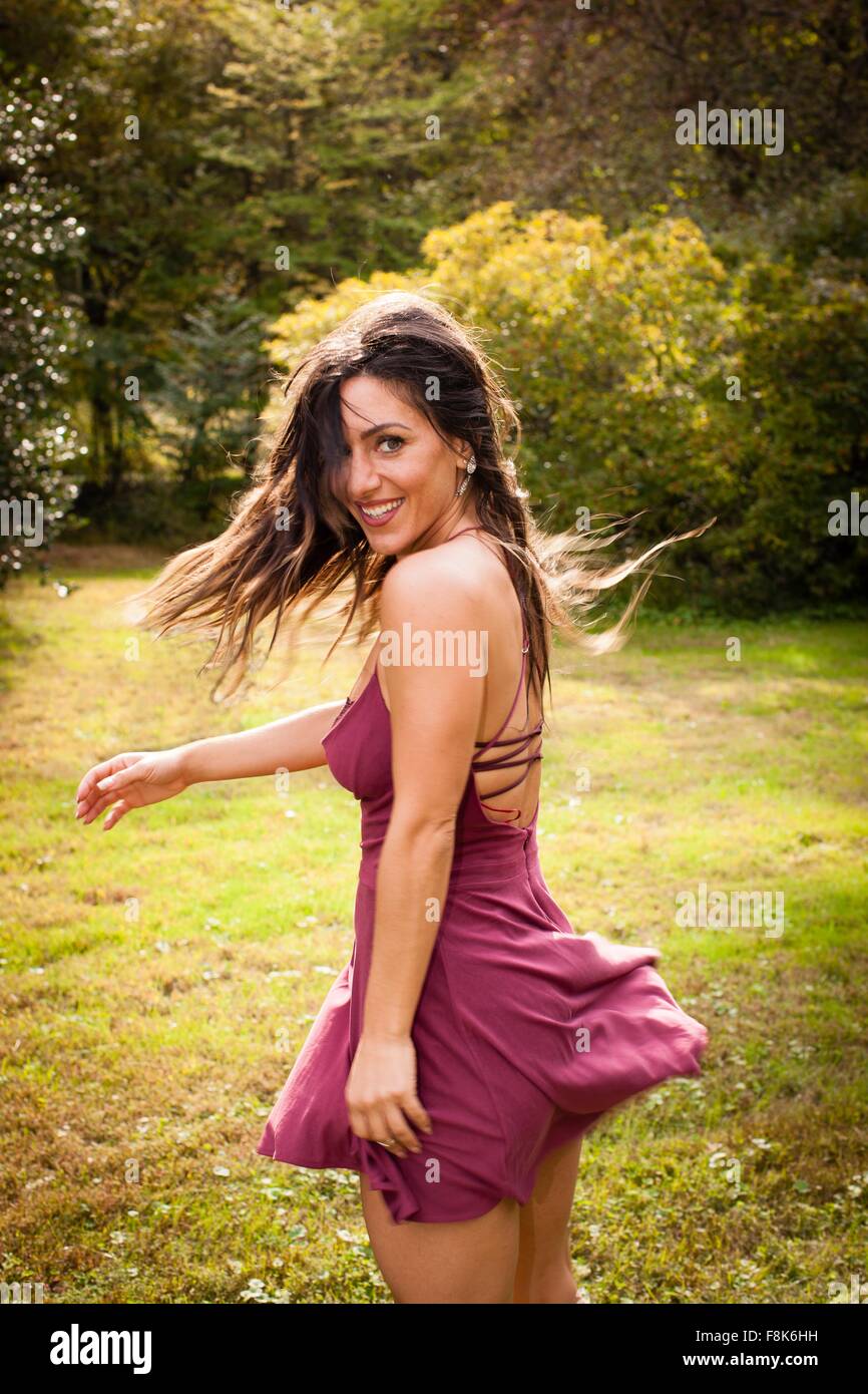 Mujer adulta media luciendo vestidos cortos bailando en el pasto, mirando por encima del hombro a la cámara sonriendo Foto de stock
