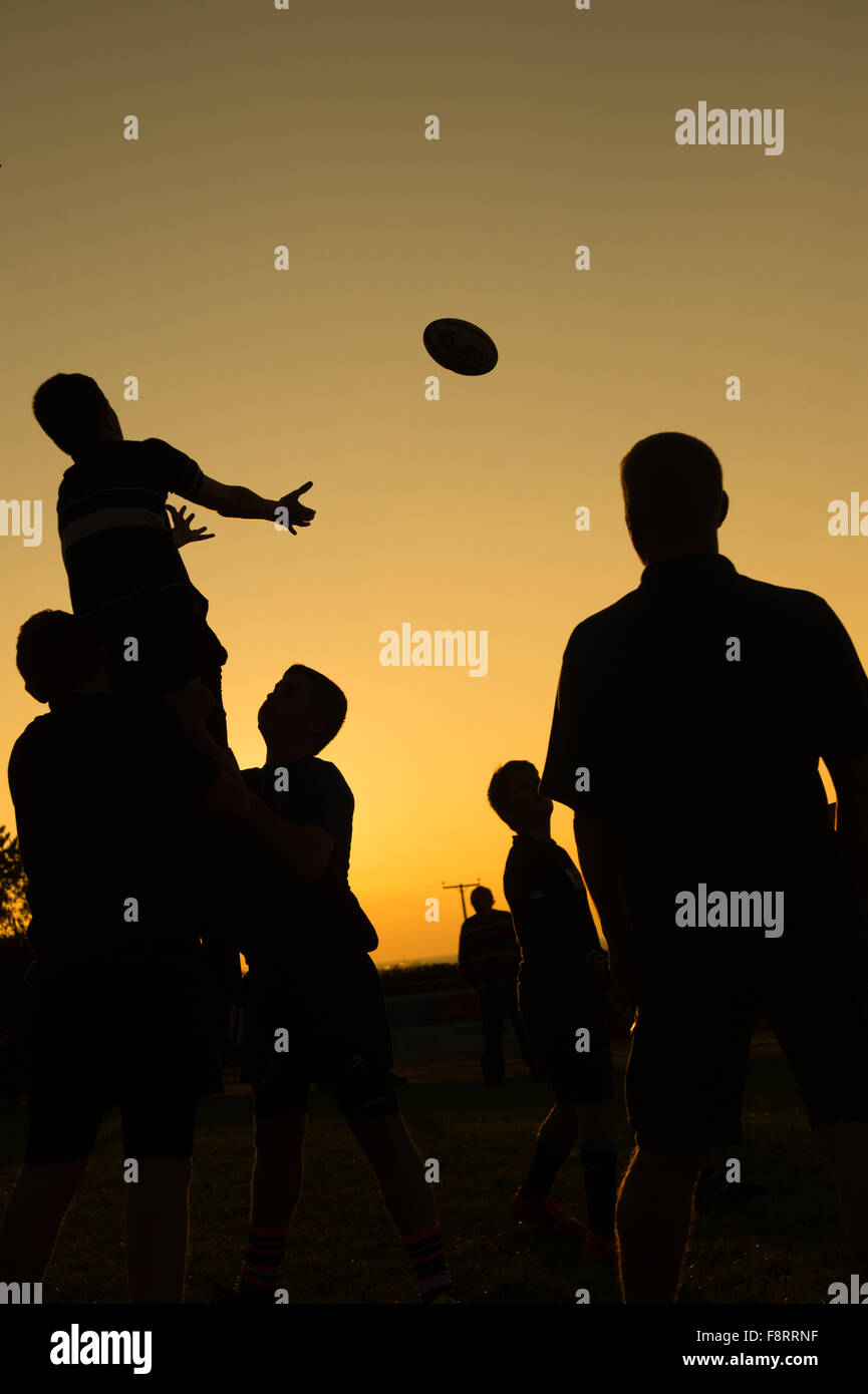 Siluetas de hombres adolescentes niños toman parte en rugby formación coaching practicando la manipulación de bolas salida de línea, al atardecer, Aberaeron Gales UK Foto de stock