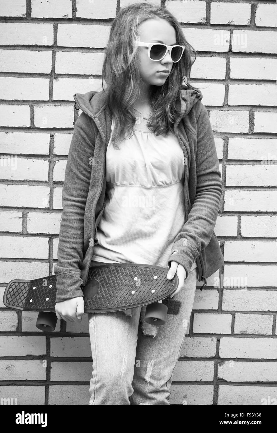 Adolescente en jeans y gafas de sol posee monopatín sobre urban pared de ladrillo, fotos en monocromo de fondo Foto de stock