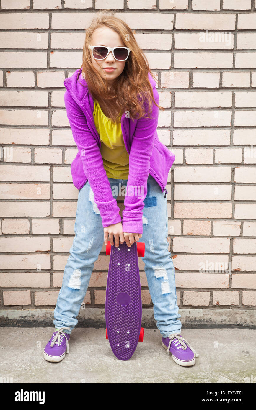 Rubia adolescente en jeans, gafas de sol y ropa deportiva mantiene cerca de monopatín urbano gris pared de ladrillo Foto de stock