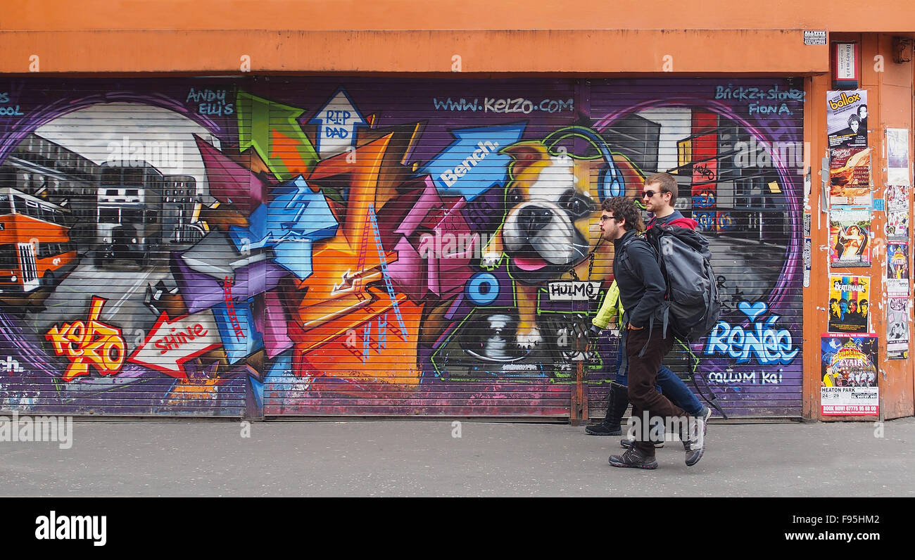 3 hombres jóvenes caminando pasado algunos street art en persianas en Stevenson Square, Lever Street, el centro de la ciudad de Manchester, Reino Unido. Foto de stock