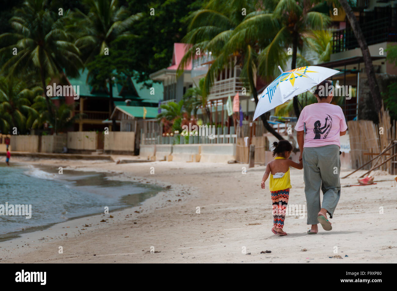 Madre e hija caminando por la playa de arena blanca con sombrilla Foto de stock
