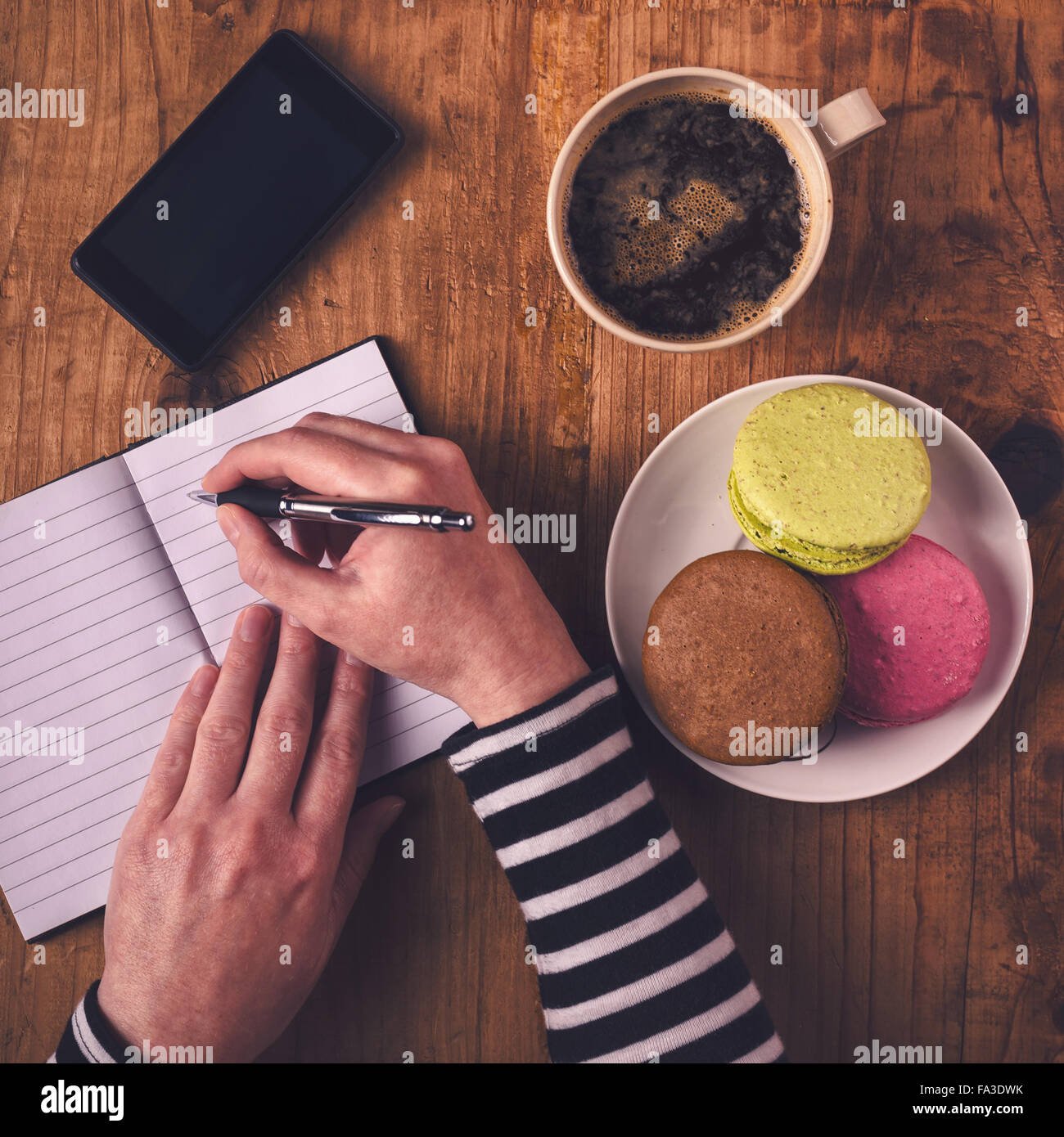 Mujer escribiendo en el portátil durante el desayuno, vista superior de manos femeninas con lápiz, taza de café, teléfono móvil y macaron cookie Foto de stock