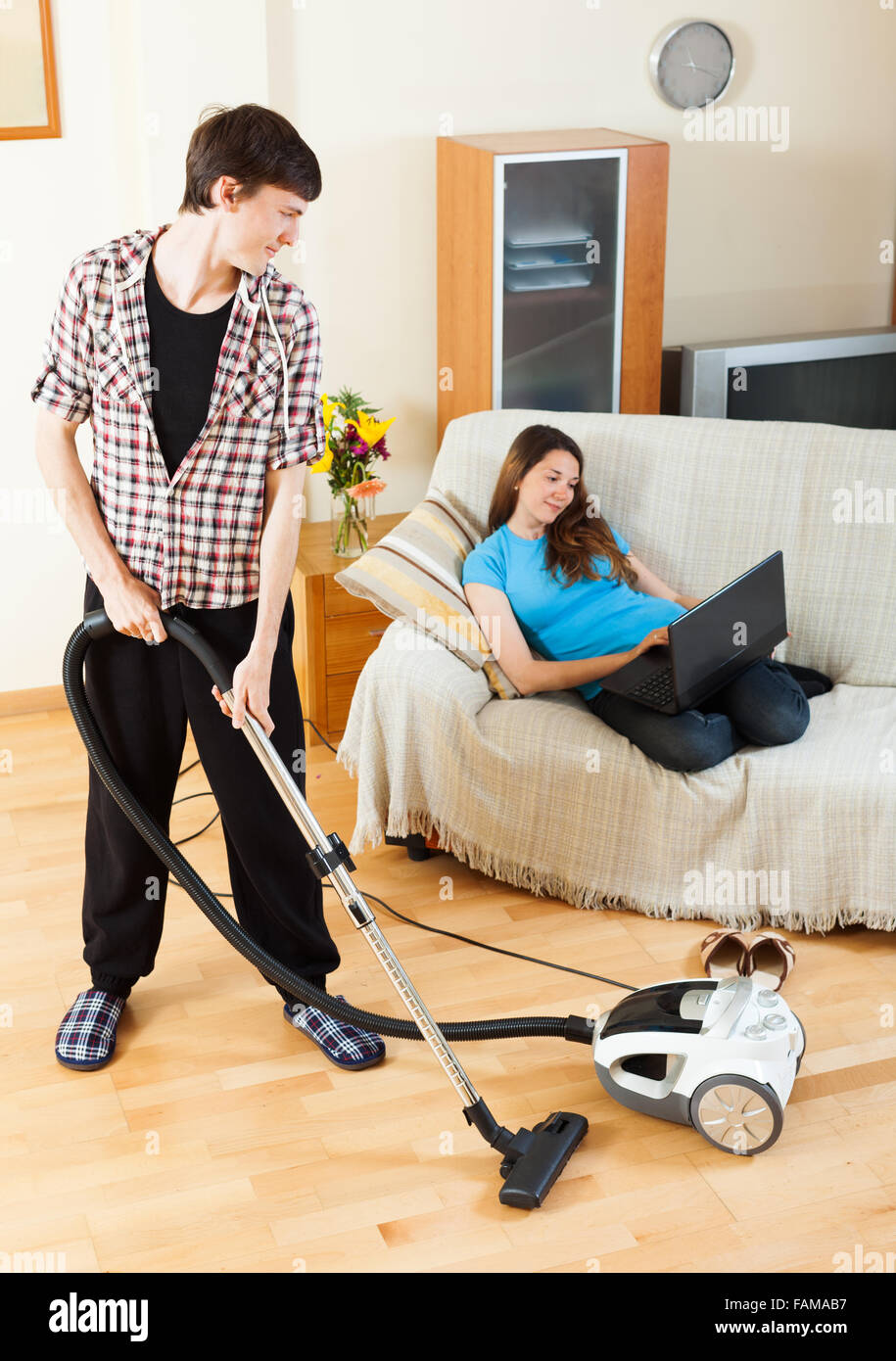 Hombre limpiar con aspiradora piso mientras su esposa acostada con portátil  de sofá en casa Fotografía de stock - Alamy