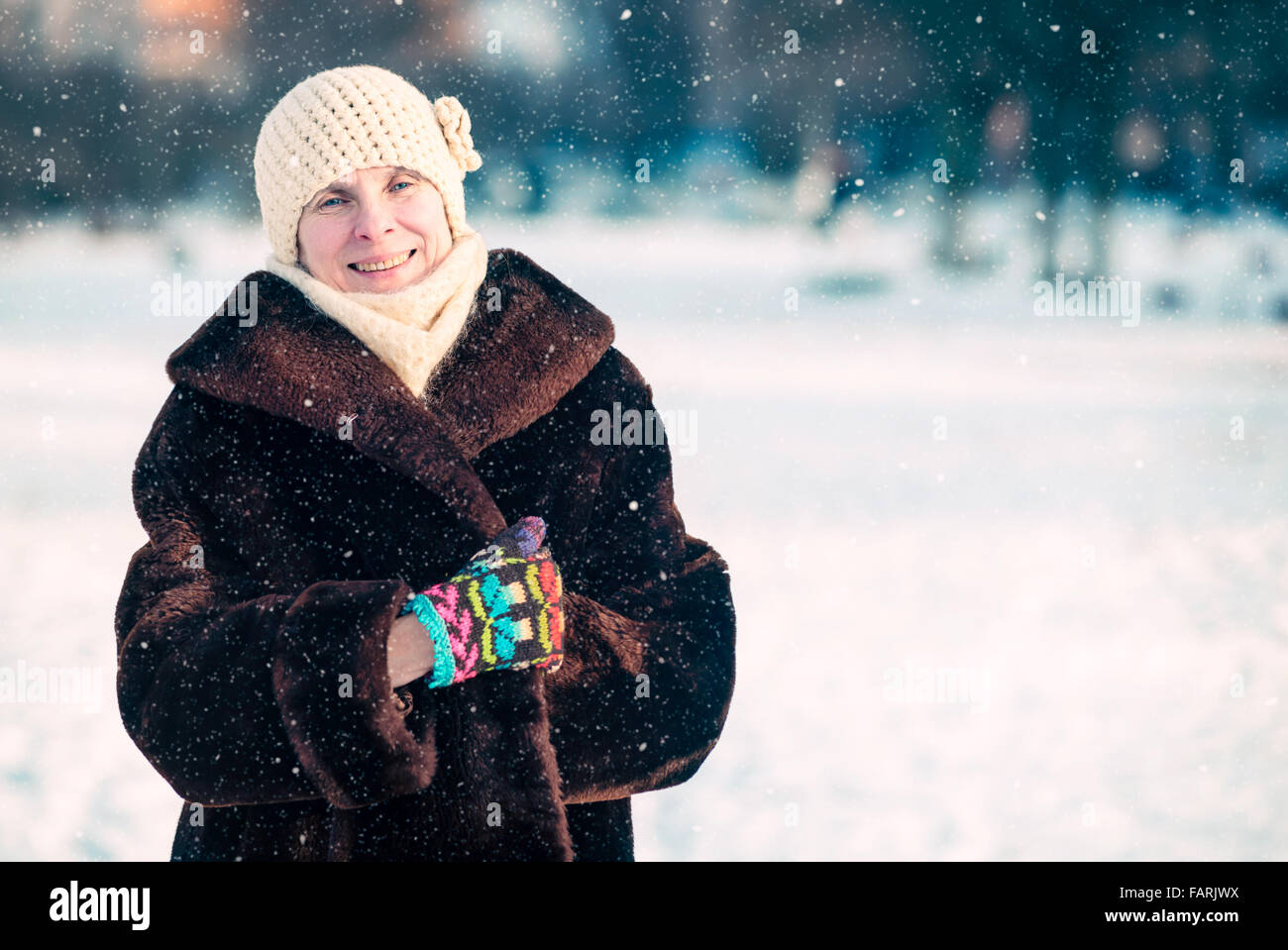 Un invierno retrato de una sonriente mujer adulta senior llevar gorro de lana, bufanda y guantes de color, con un fondo de nieve Foto de stock