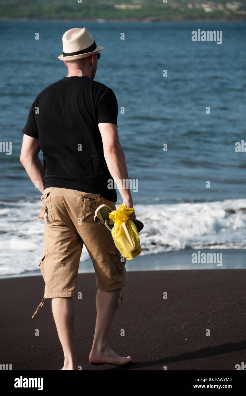 Hombre con camisa negra y sombrero blanco caminar sobre la playa de arena negra en la costa Foto de stock