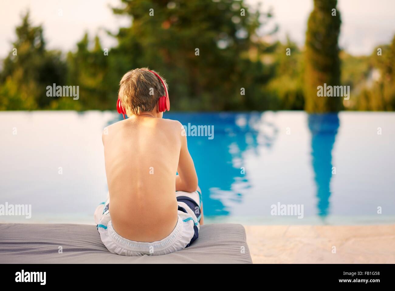 Vista trasera del adolescente usando audífonos sentado en piscina infinity Foto de stock