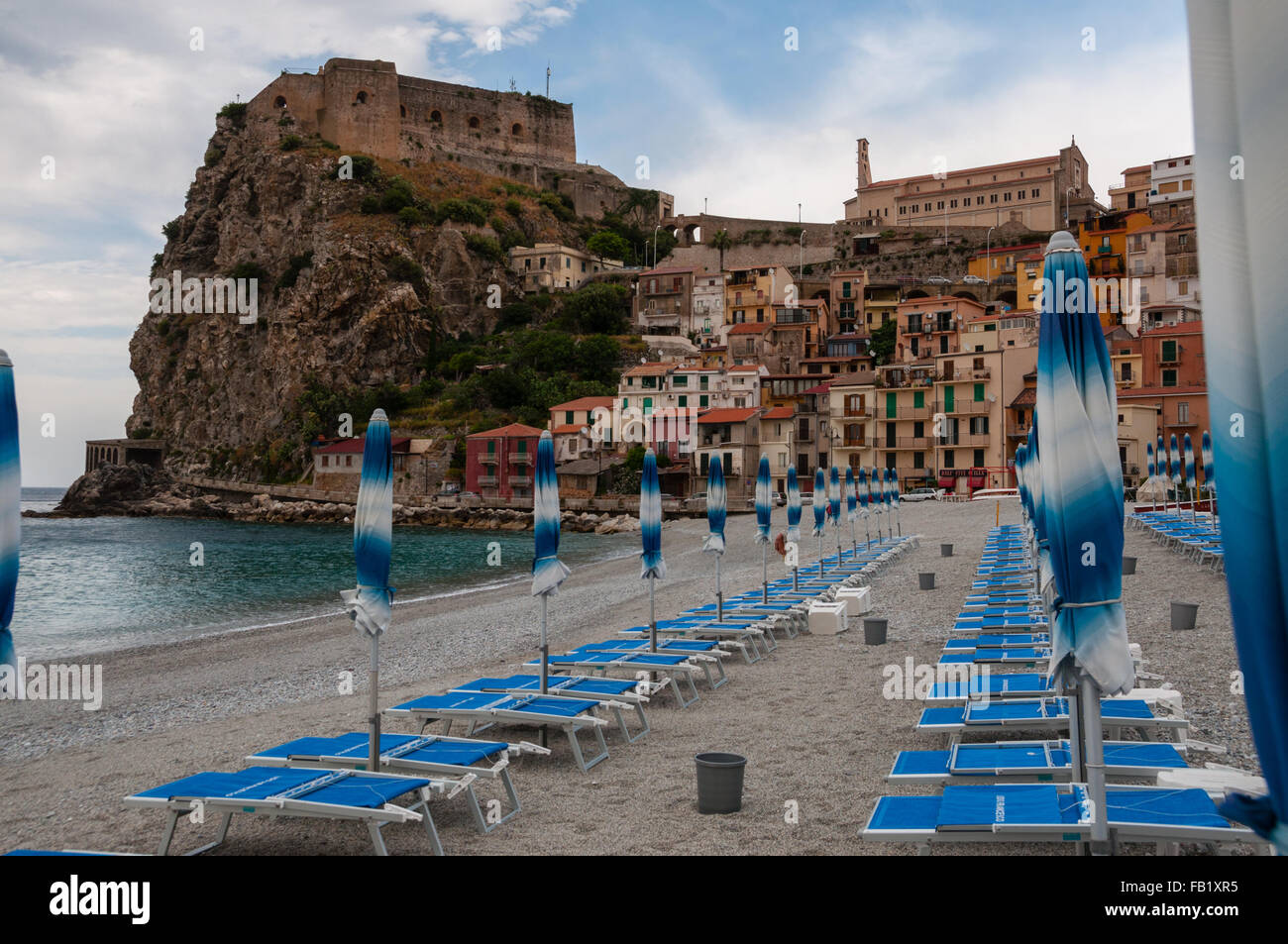 Paraguas y beachchair azul en la playa en frente de la pequeña ciudad italiana sobre un acantilado bajo un cielo azul Foto de stock