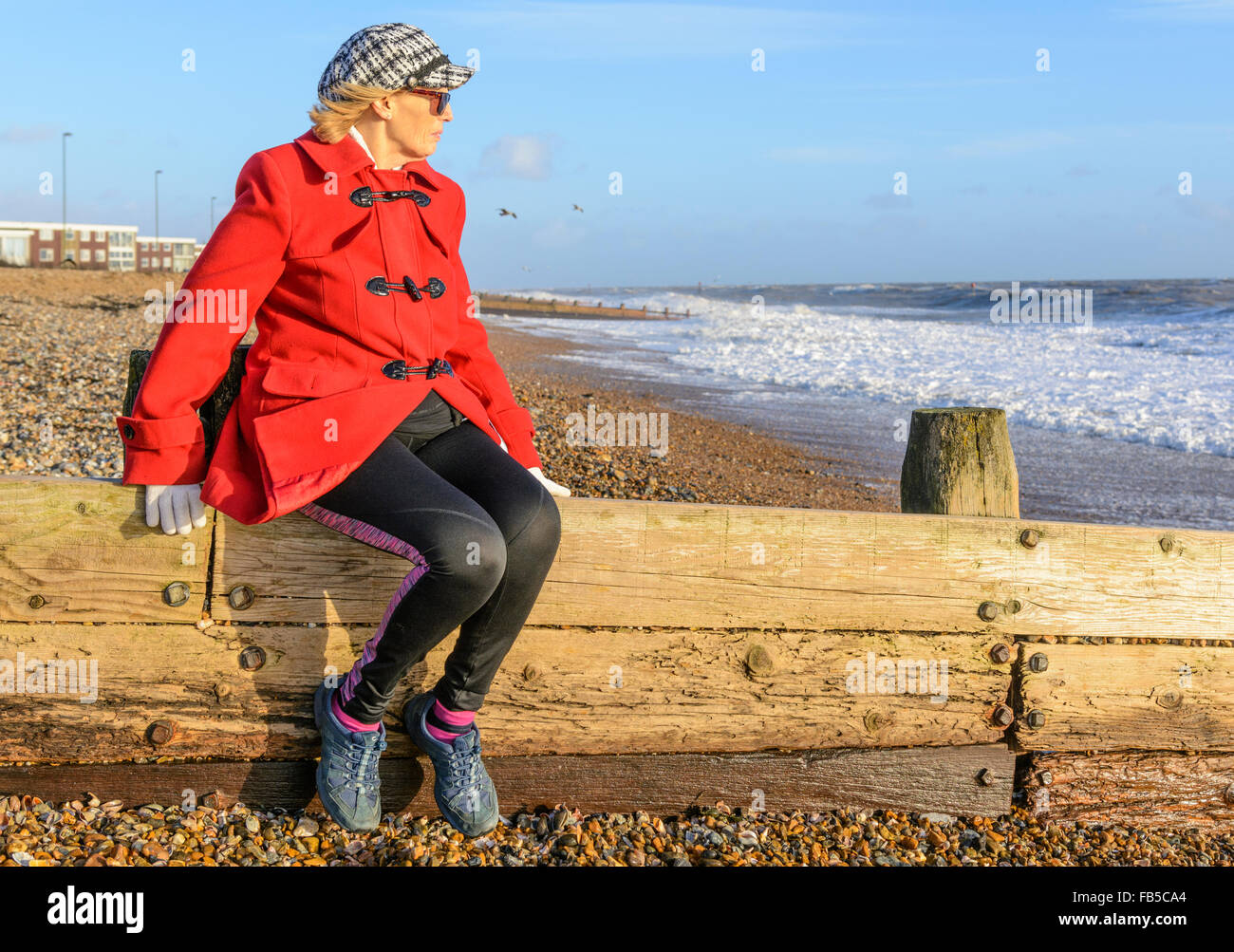 Señora de mediana edad sentado en un espigón en la playa en invierno, mirando al mar en un día frío en West Sussex, Inglaterra, Reino Unido. Foto de stock