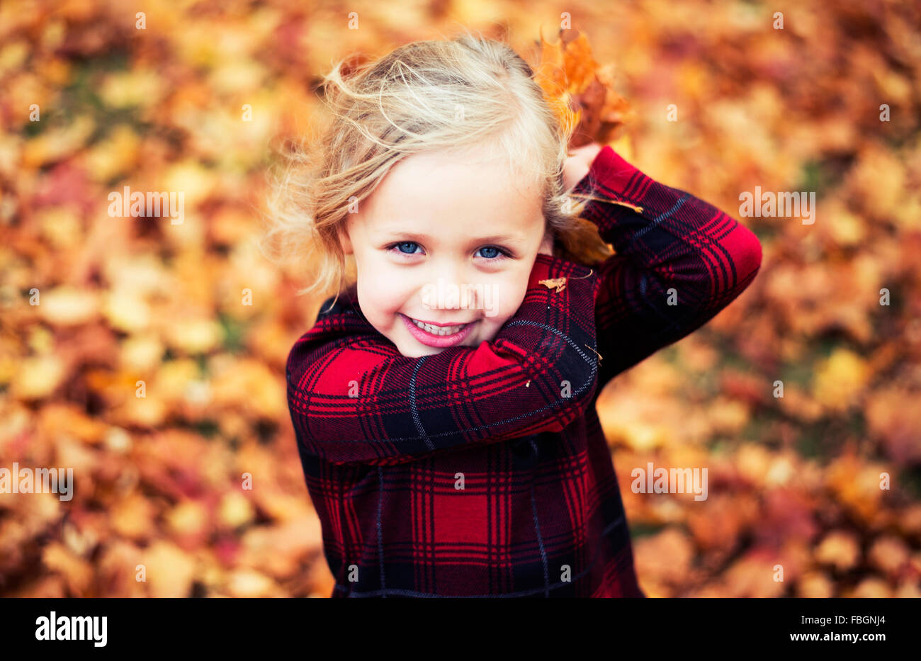 Linda chica con el pelo rubio ojos azules y rojos y negros camisa marcada a arrojar hojas de naranja en otoño Foto de stock