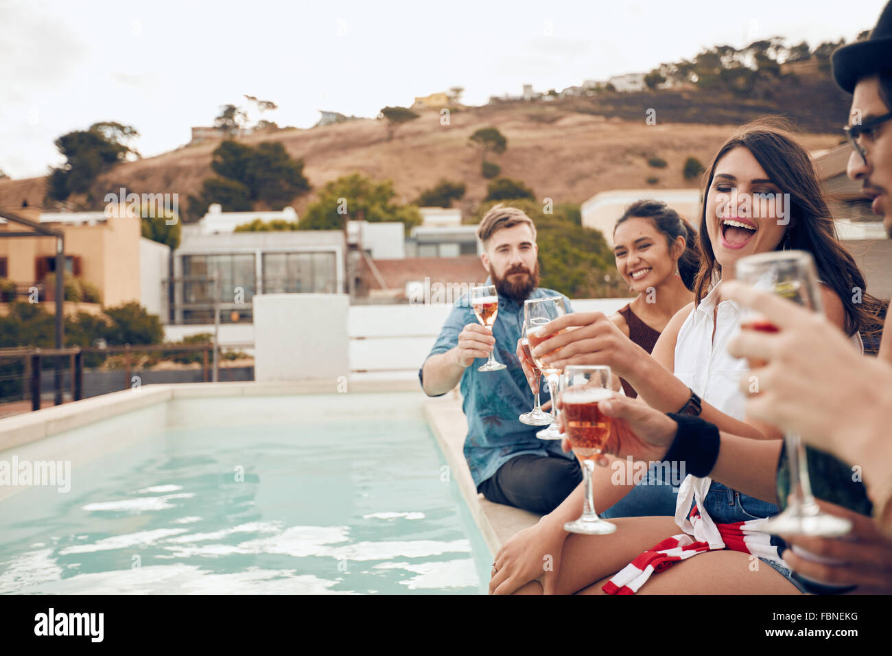 Disparó al aire libre de feliz grupo de jóvenes sentados en el borde de la piscina para beber vino. Amigos disfrutando multirracial y toas Foto de stock