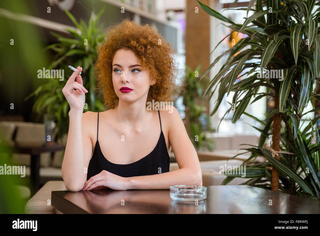 Pensativo pelirroja mujer con cabello rizado fumar en restaurante. Foto de stock