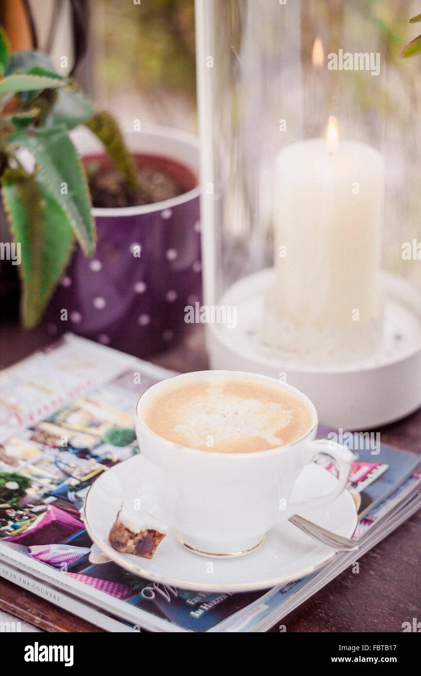 Tabla con una taza de café, dulces, revistas, linterna con velas encendidas y planta de casa Foto de stock