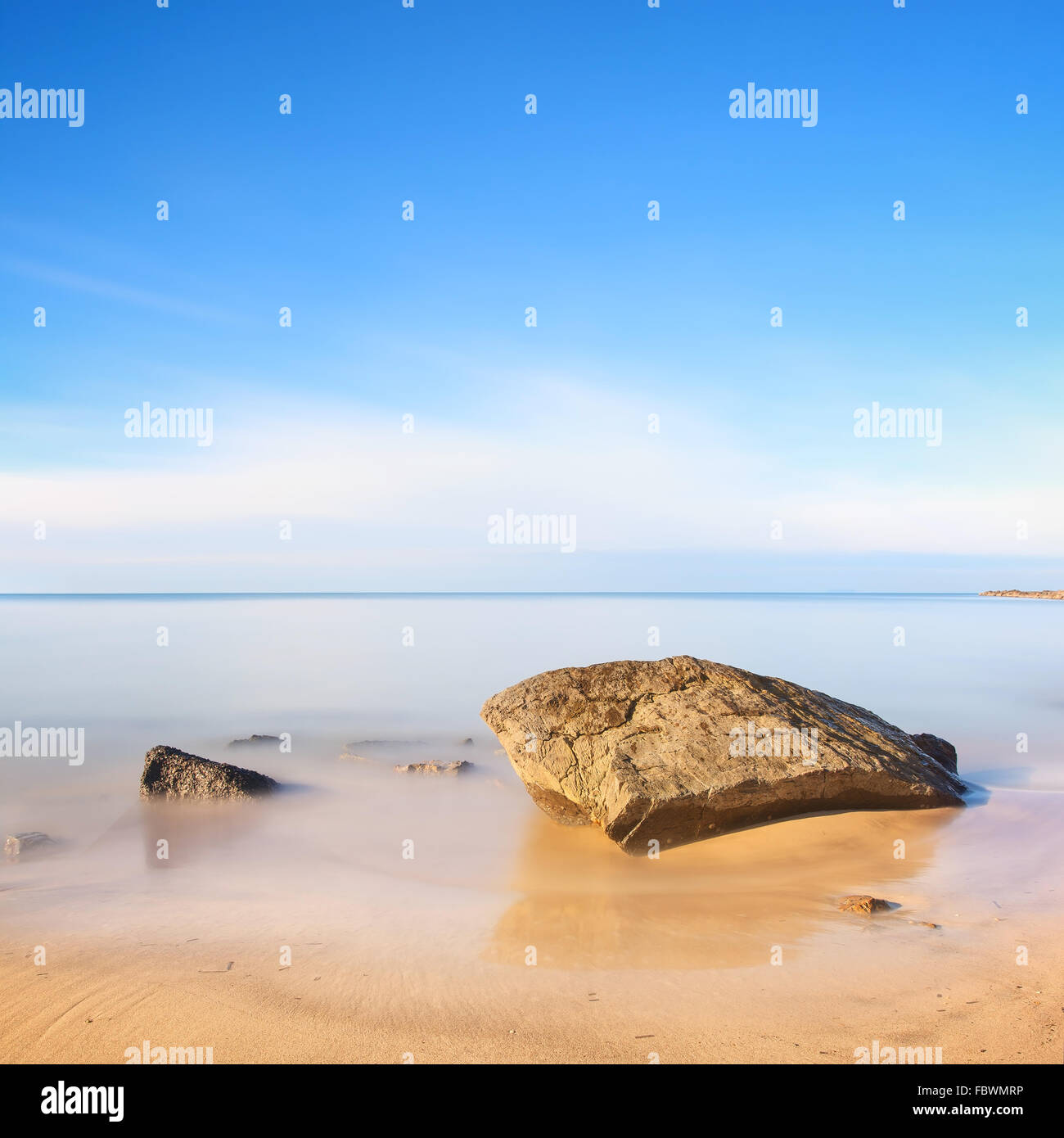 Una roca plana sobre una playa de arena dorada y azul océano. Fotografía de larga exposición. Foto de stock