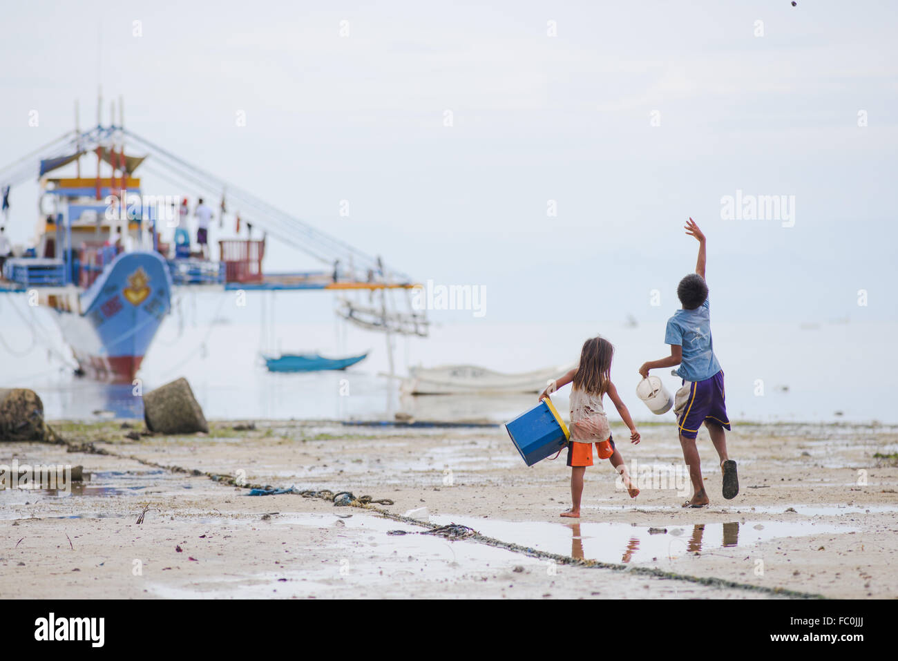 Dos niños jugando, arrojando piedras en el mar con un barco pesquero con estabilizadores Foto de stock