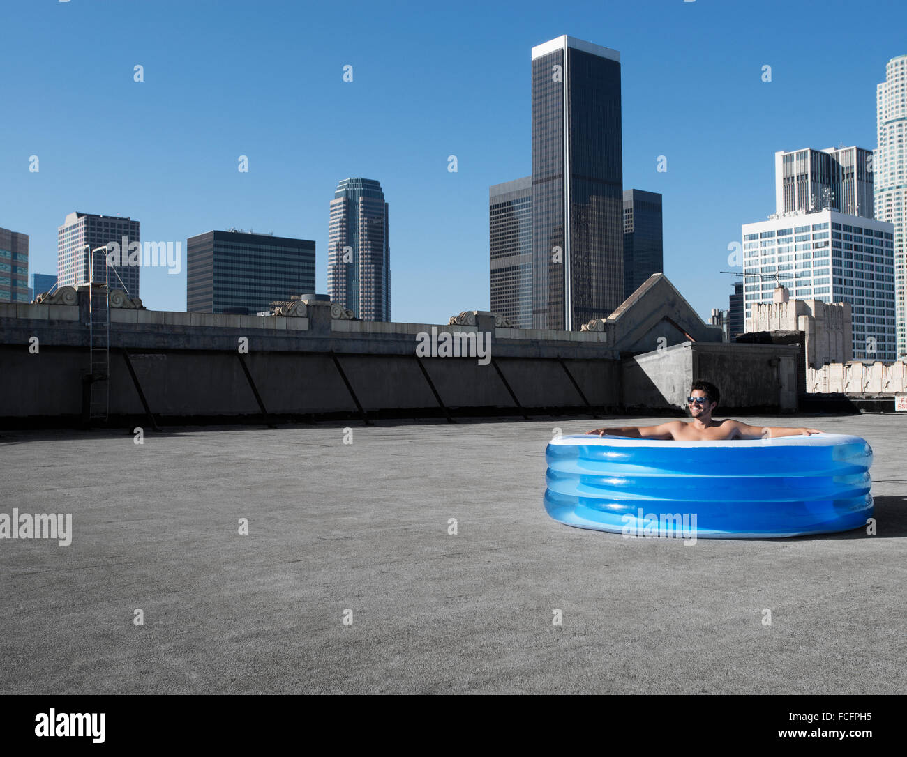Un hombre sentado en una pequeña piscina de agua inflable en una azotea de la ciudad, enfriando. Foto de stock