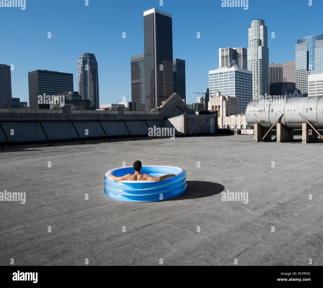 Un hombre sentado en una pequeña piscina de agua inflable en una azotea de la ciudad, enfriando. Foto de stock