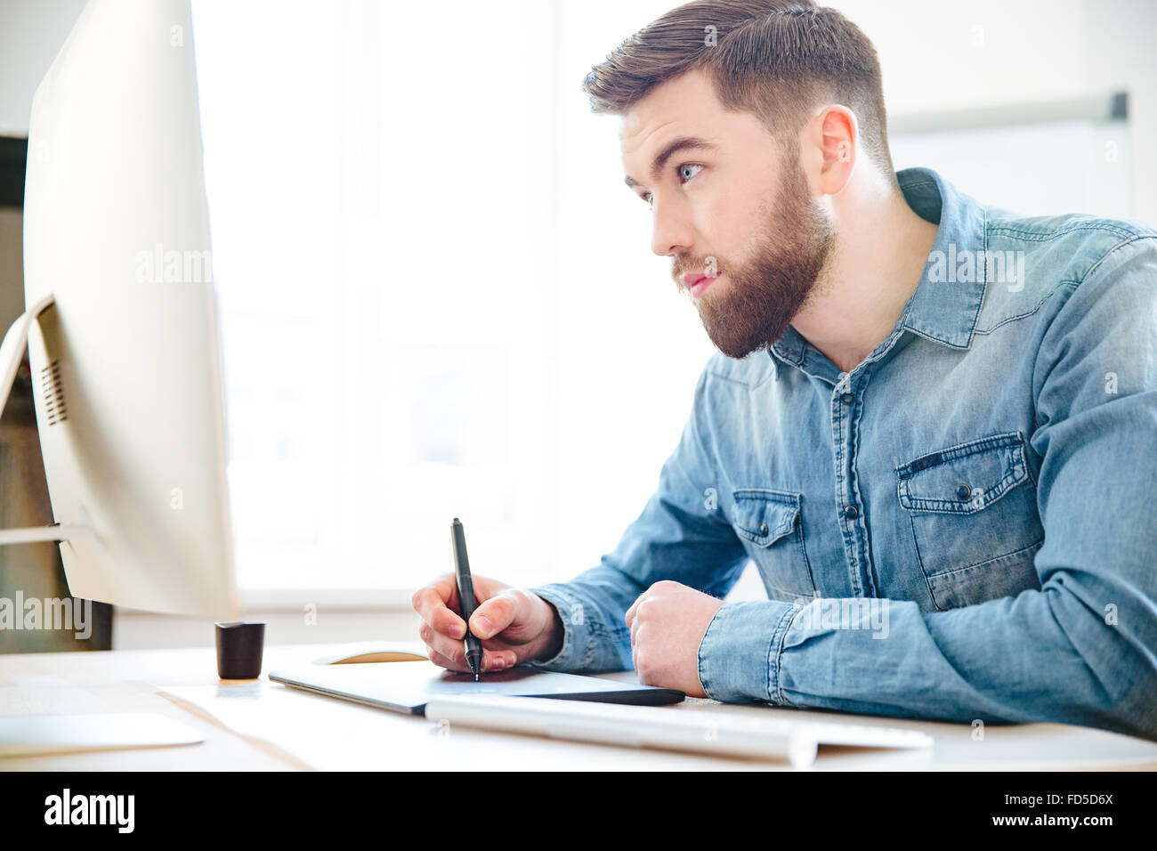 Concentrado apuesto joven diseñador con barba en camisa azul dibujo con ordenador y tableta gráfica en la oficina Foto de stock