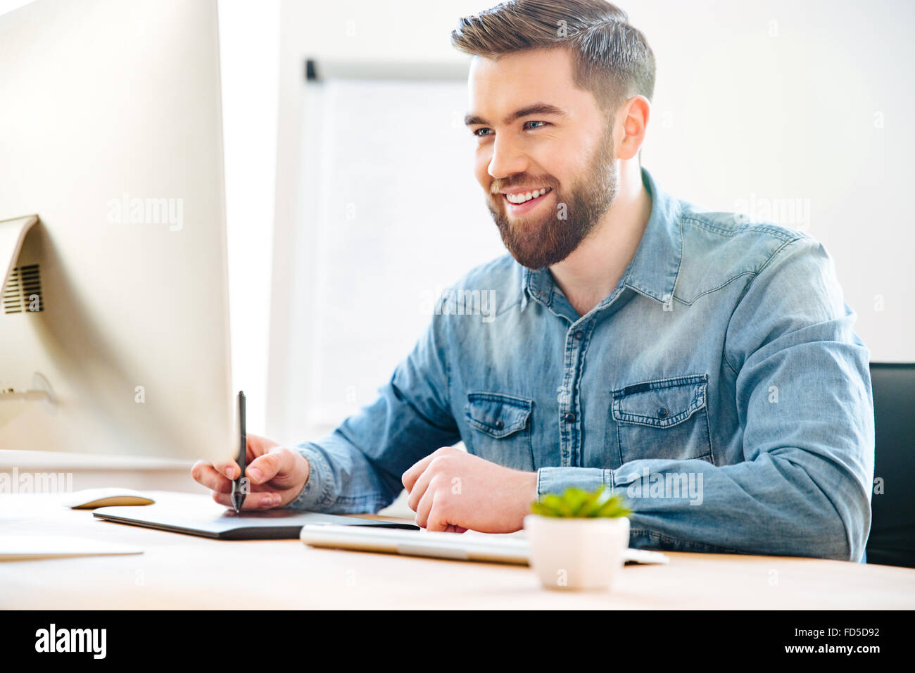 Feliz atractivo diseñador joven sentado en la oficina y el dibujo sobre la tableta gráfica Foto de stock