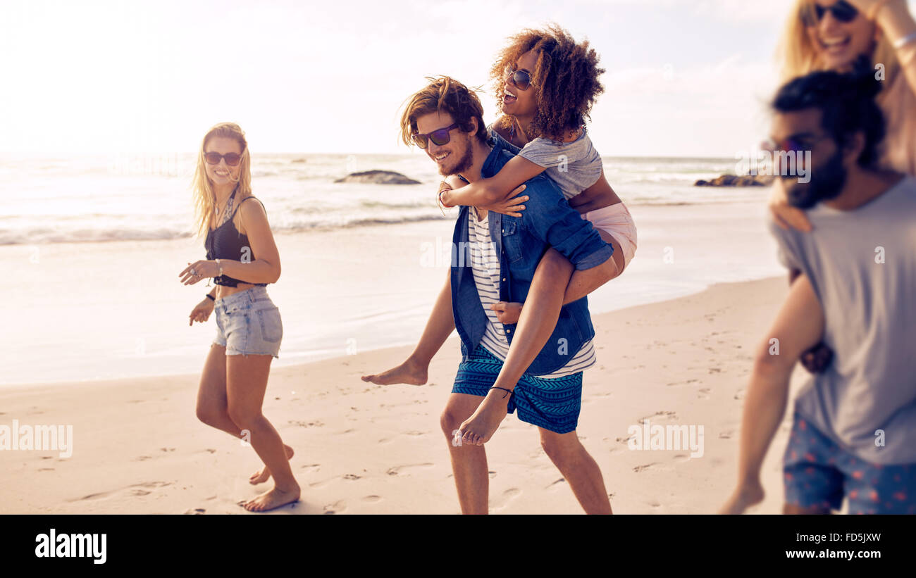 Dos hombres jóvenes felices dando sus novias piggyback cabalgatas en la playa. Grupo de amigos disfrutando de unas vacaciones en la playa. Foto de stock