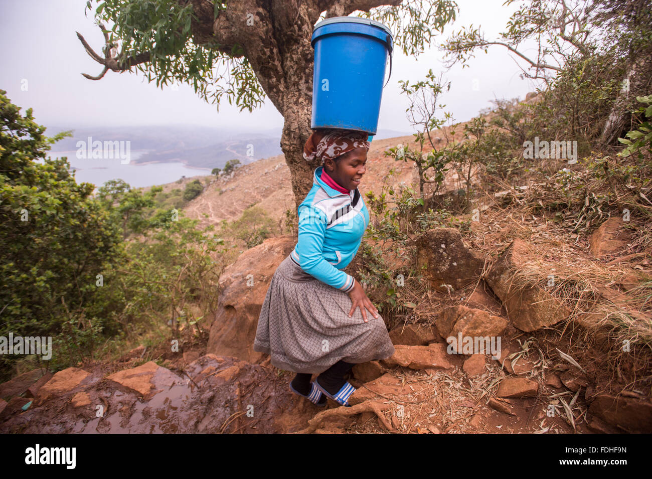 Mujer que llevaba un balde sobre la cabeza sobre el terreno rocoso en la región de Hhohho de Swazilandia, en África. Foto de stock