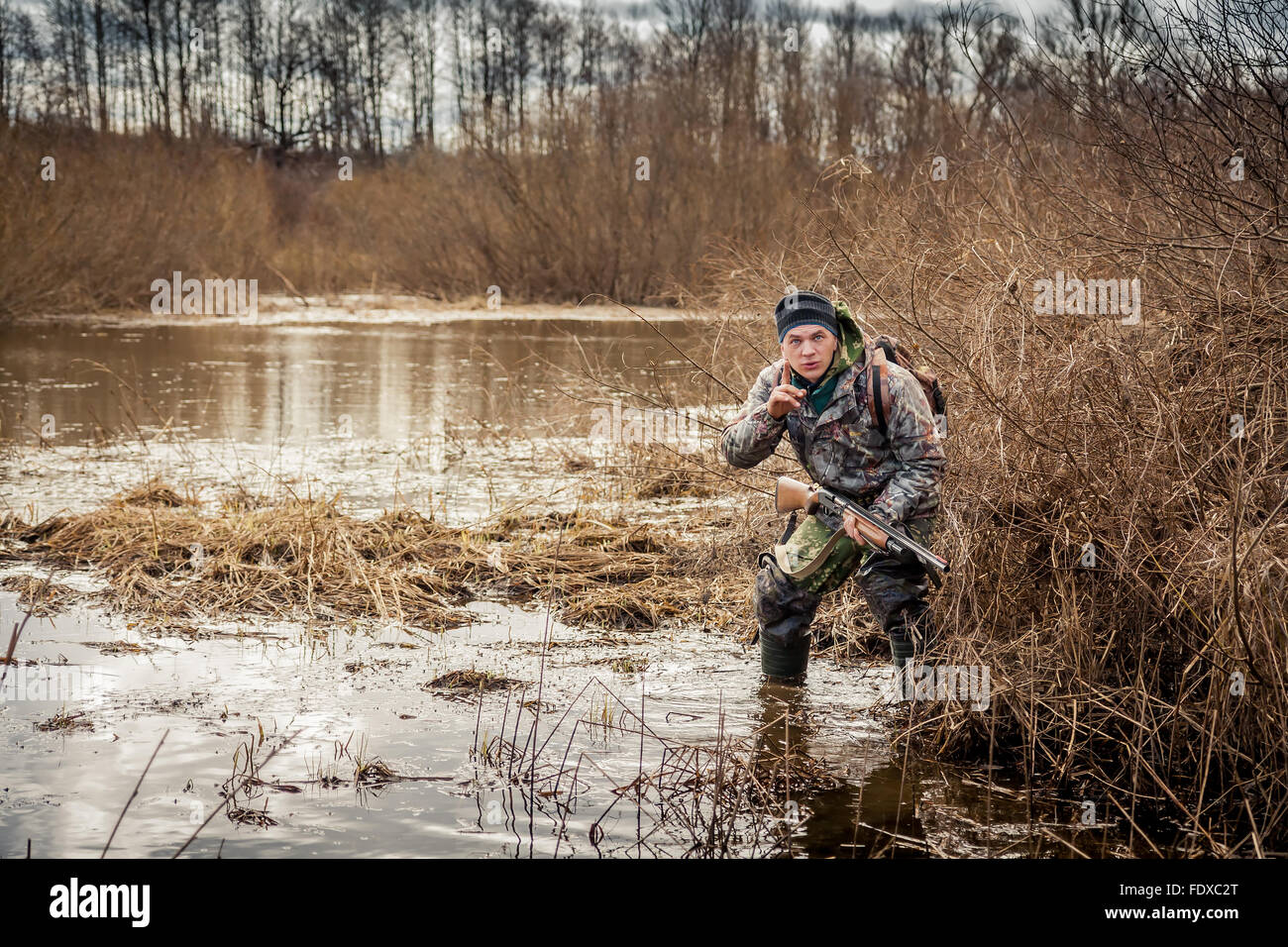 Hunter hombre vadeando a través del pantano que acechan en los arbustos y muestra un gesto para estar tranquilo Foto de stock