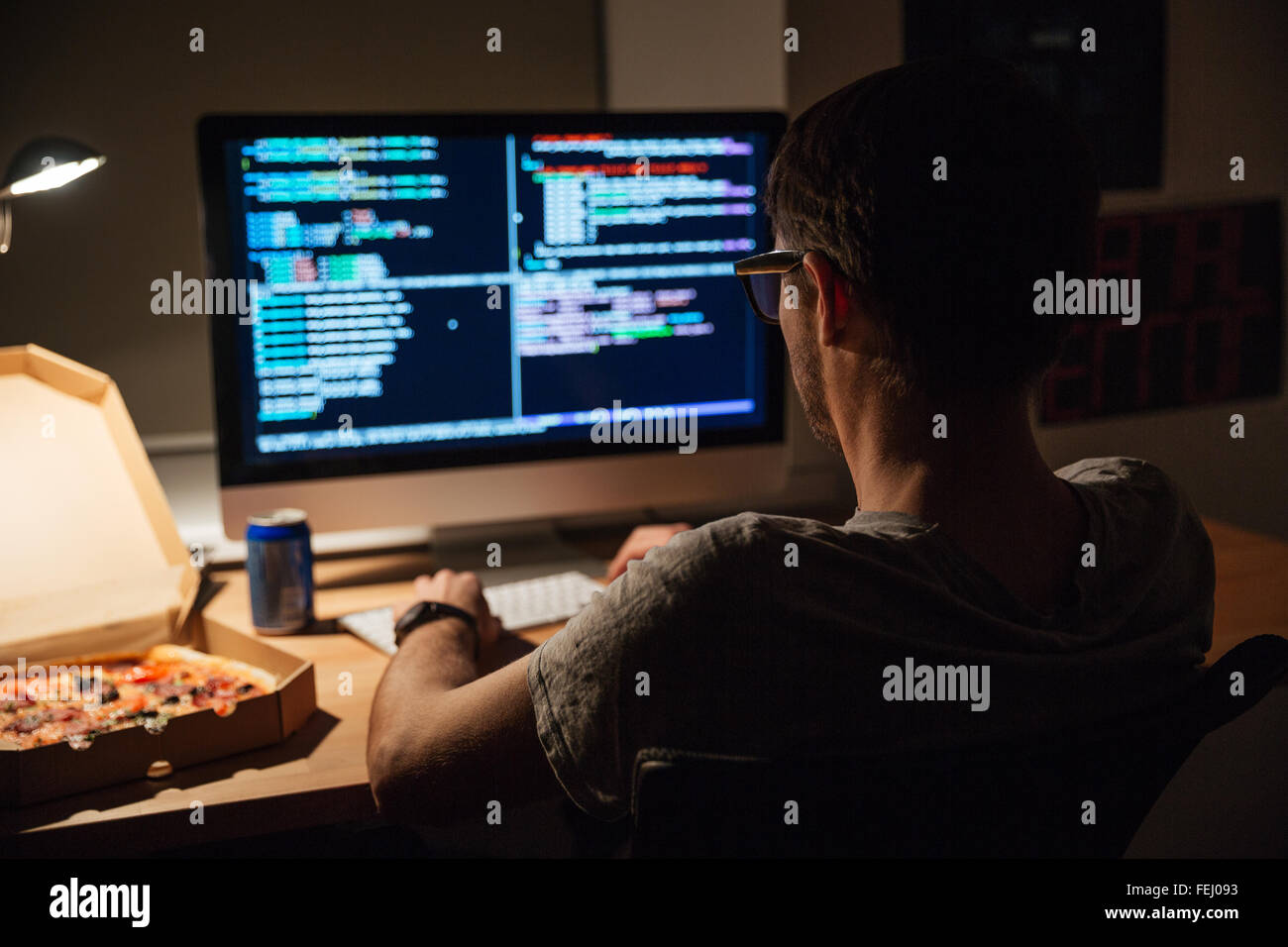 Vista posterior del programador joven centrado en vidrios de escribir código y comer pizza Foto de stock