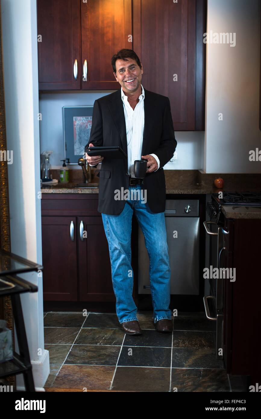 Mature hombre vestido con traje chaqueta y pantalones vaqueros en cocina,  celebración y tableta digital taza de café, mirando a la cámara sonriendo  Fotografía de stock - Alamy