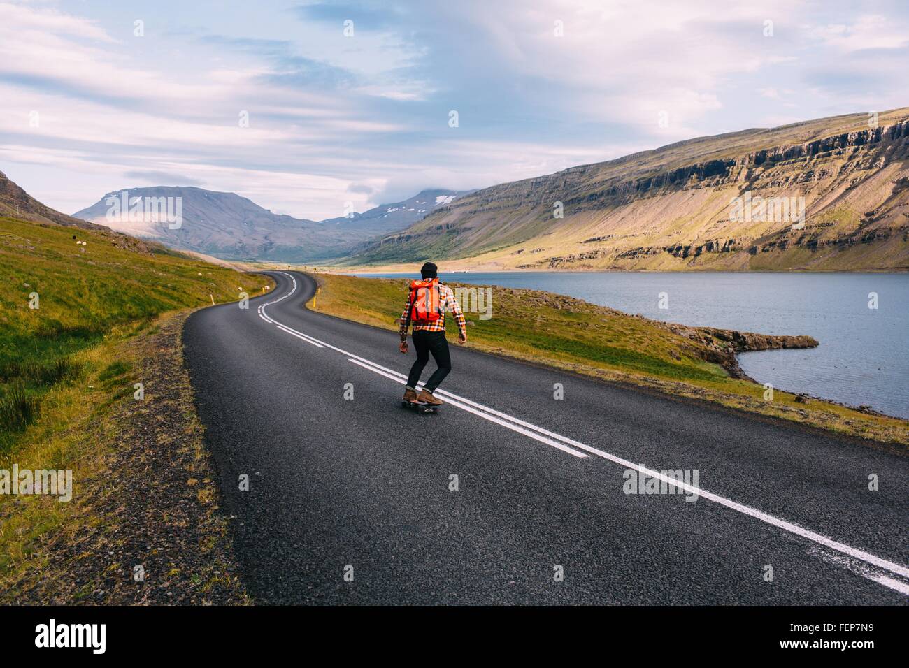 Vista trasera de mediados del hombre adulto skateboarding en curva camino abierto por el lago, Islandia Foto de stock