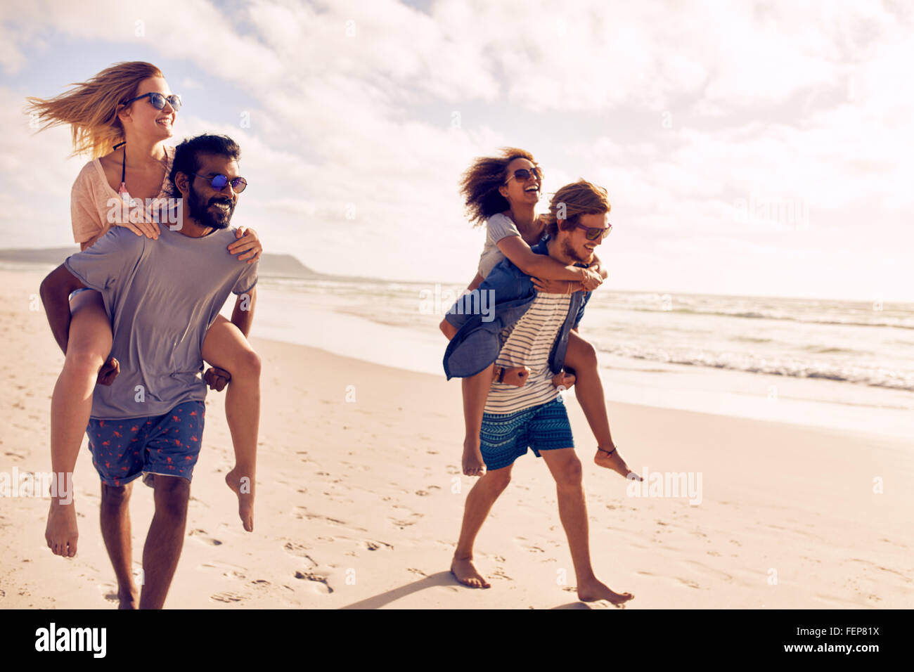 Dos hermosas jóvenes parejas caminando por la playa, con hombres que llevaban a sus mujeres en sus espaldas. Las parejas sumarse a la orilla del mar Foto de stock