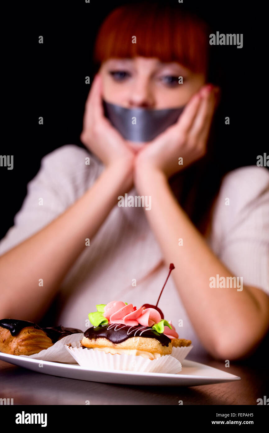 La niña, cuya boca sellada con cinta triste mirando la placa con tartas Foto de stock