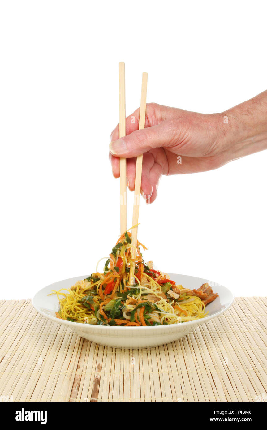 Mano sosteniendo los palillos se sumerge en un tazón de fideos de Singapur sobre un fondo blanco. Foto de stock