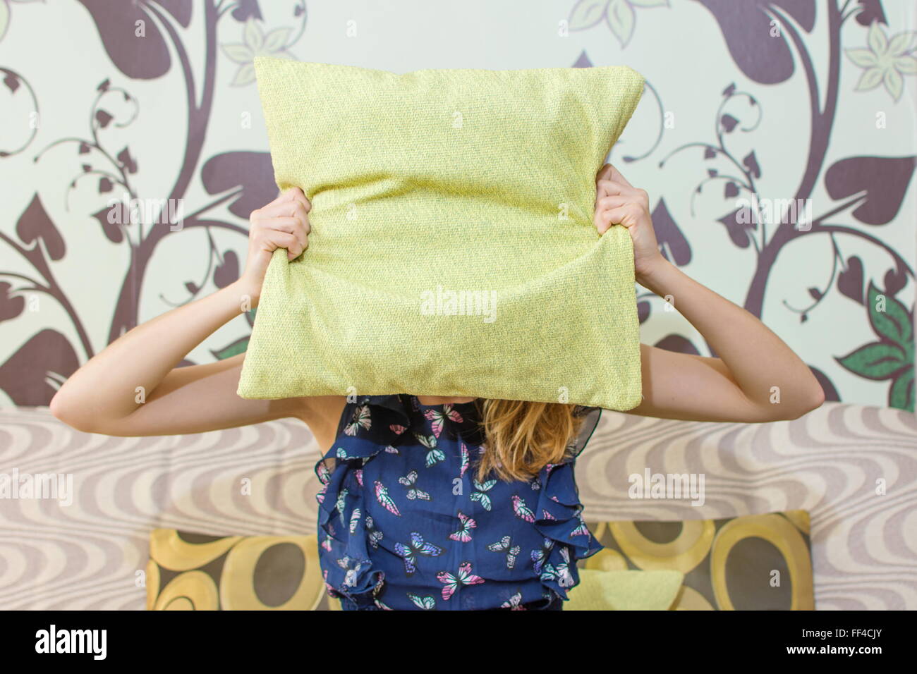Chica escondiéndose detrás de una almohada verde sentado en el sofá de la sala Foto de stock
