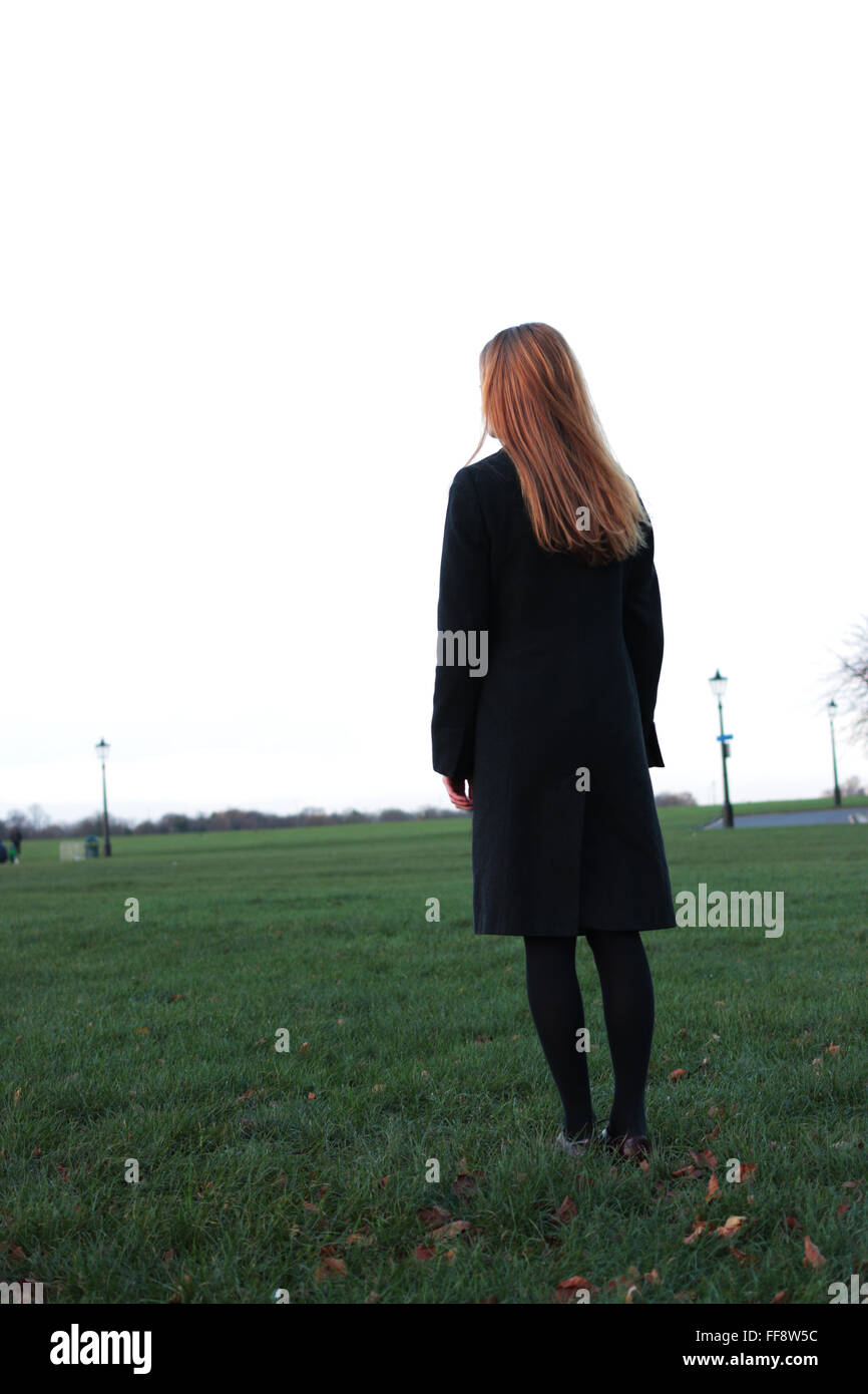 Pensativo joven mujer de pie en un parque mirando en la distancia. Foto de stock