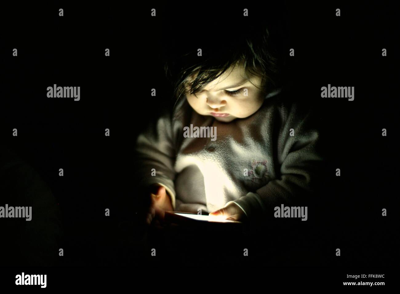 Niña de 10 meses mirando una pantalla del teléfono inteligente en una habitación oscura Foto de stock
