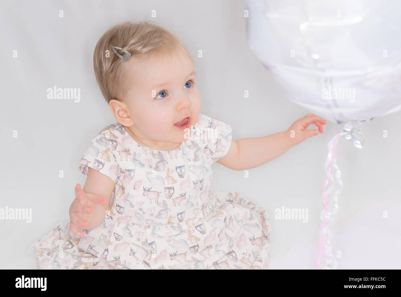 Un hermoso bebé lindo con ojos azules en un vestido delante de un fondo blanco mirando hacia un globo con un lazo atado a ella Foto de stock