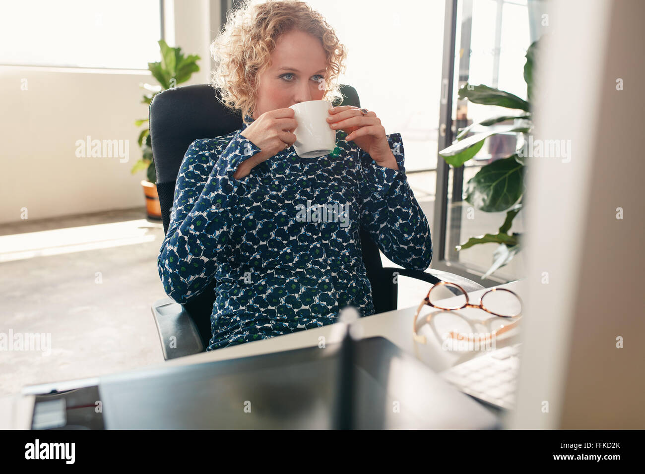 Retrato de mujer joven sentado en su pupitre bebiendo café y mirando el monitor del equipo. Diseñador reflexiva con café. Foto de stock