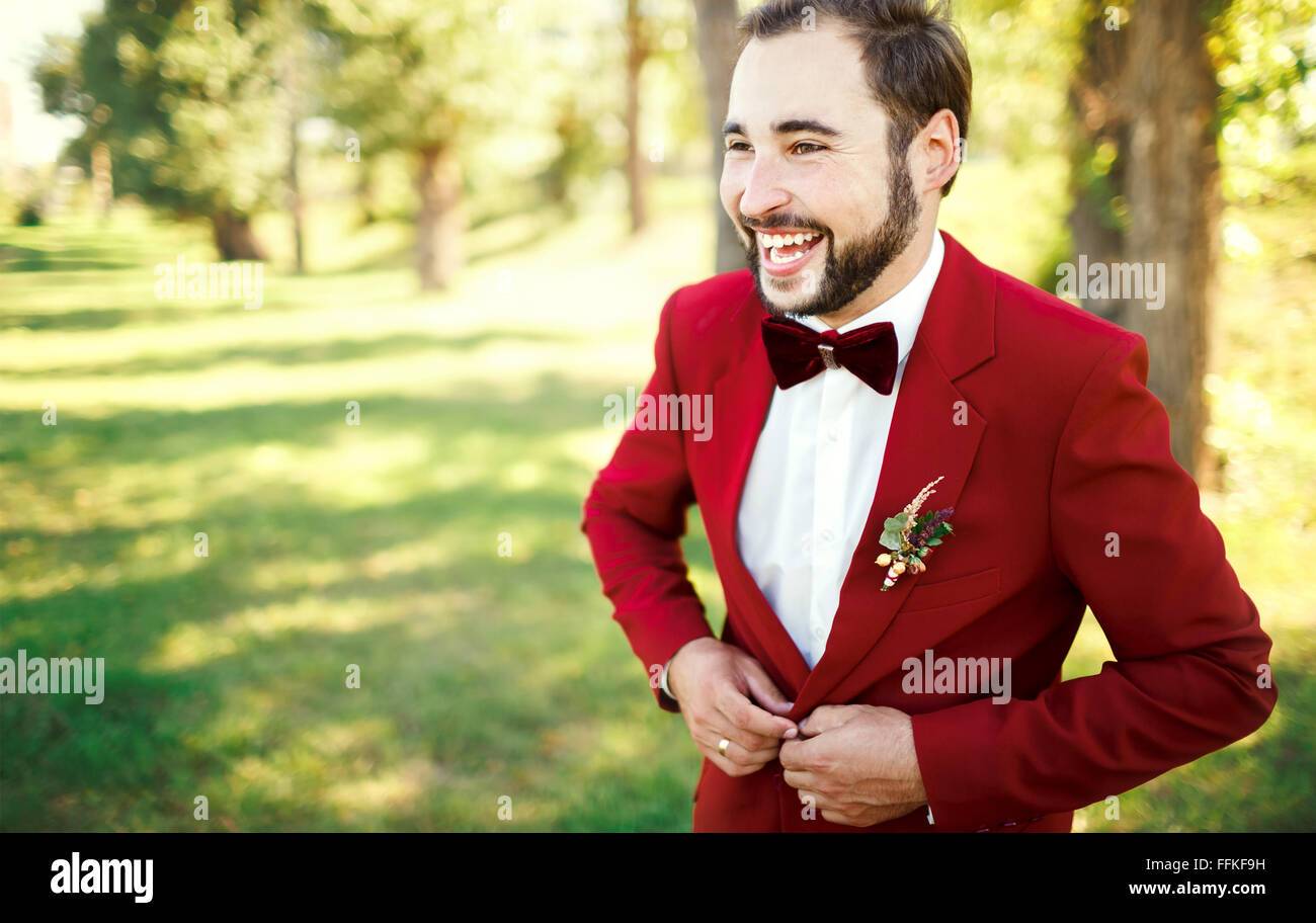 Elegante traje de novio en tuxedo risas Marsala, borgoña rojo pajarita. Hombre abotonarse la chaqueta exterior. Peinado profesional, barba, bigote. Preparativos de boda, preparándose. Espacio para copiar texto. Foto de stock