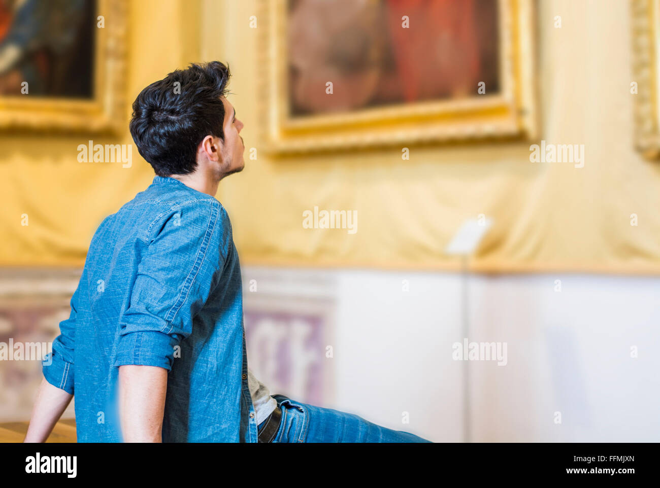 Disparo de medio cuerpo de un guapo joven reflexivo, Mirando la pintura sentado en un banco dentro de un museo Foto de stock