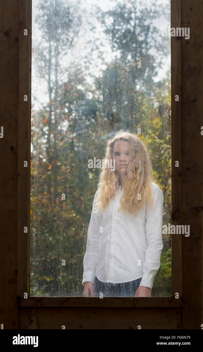Retrato de una adolescente caucásicos con largo cabello rizado rubio de pie en el otro lado de una ventana sucia enmarcado por la madera antigua Foto de stock