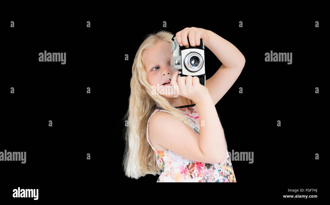 Joven con largo pelo rubio tomando una fotografía con una cámara vintage de pie contra un fondo negro Foto de stock