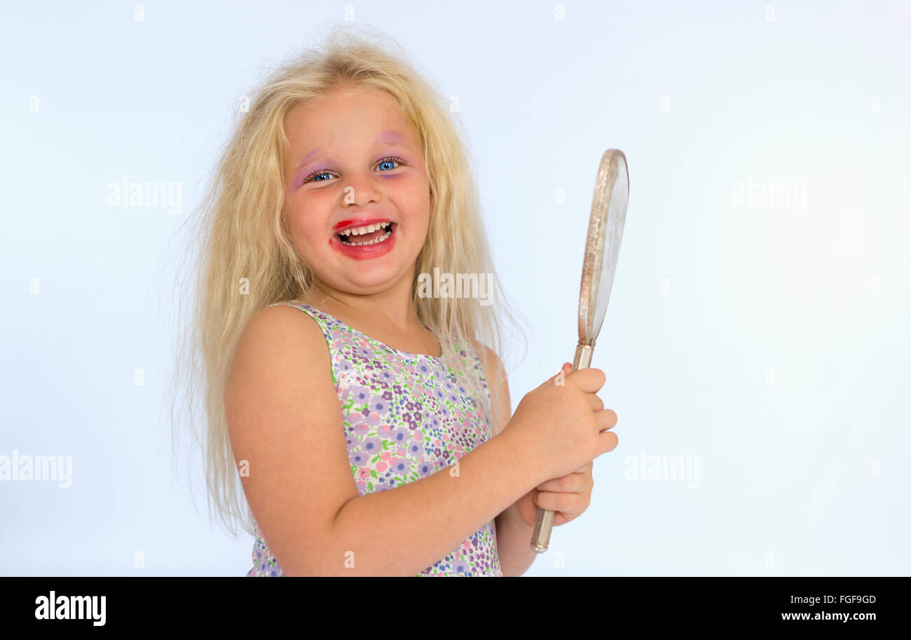 Muchacha con cabello rubio vistiendo maquillaje manchado sosteniendo un espejo, riendo Foto de stock